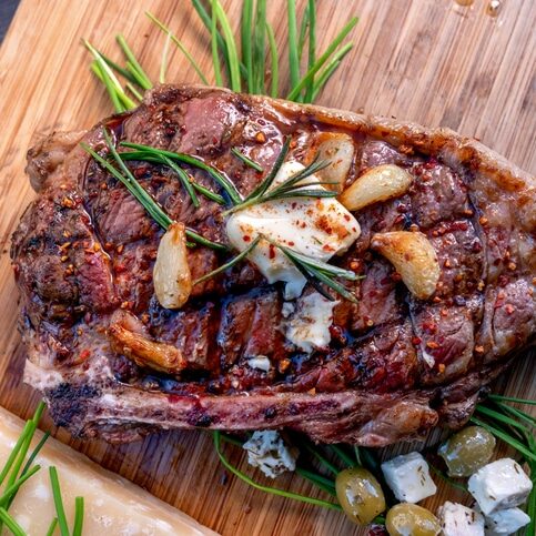 Ribeye Steak Just Off A Charcoal Grill Resting On A Wood Cutting Board, Seasoned With Rosemary, Garlic Cloves, Butter, Cheese, Olives, And Paprika