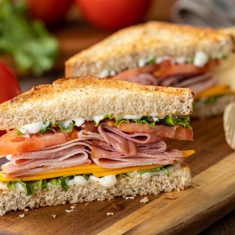 Ham sandwich with cheese, lettuce and tomato on whole grain bread.  Lettuce and tomatoes in background